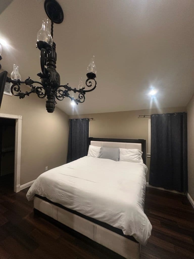 Modern bedroom with a king-size bed, elegant chandelier, and dark curtains on wooden floors.